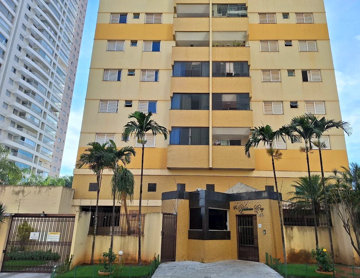 Frente do prédio com a portaria de granito preto, o portão de pedestres, o portão da garagem à esquerda e jardins com palmeiras.